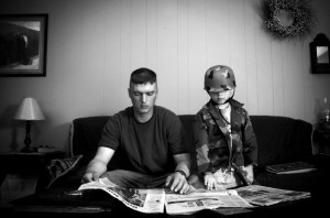 A father home home on leave reading the newspaper with his son, who wears his dad’s helmet.